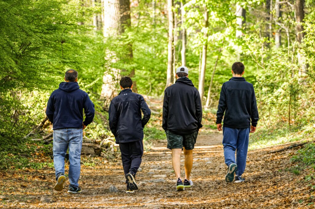 Four men in casual attire walking on a sunlit forest trail, enjoying nature.